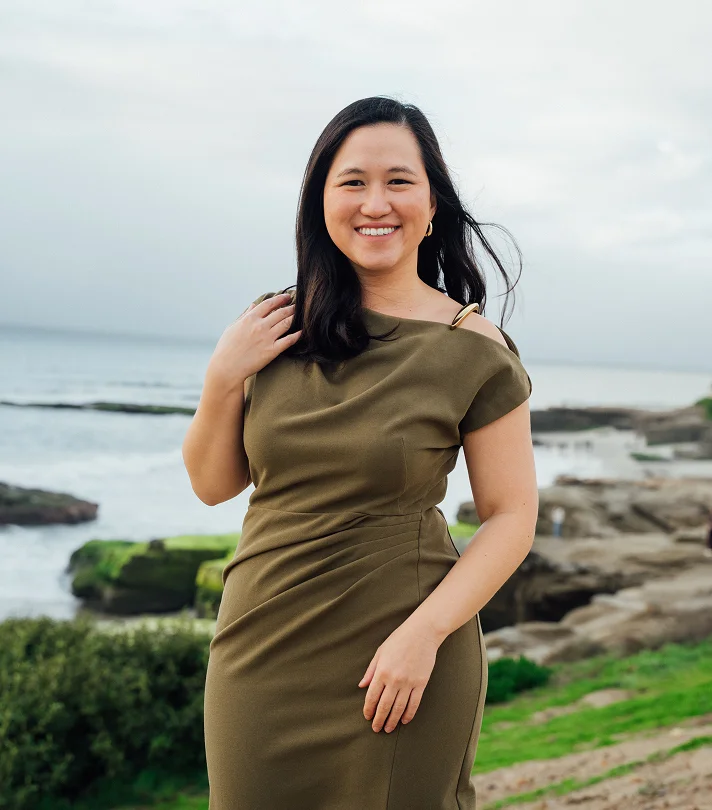 Doctor Tiffany Pham MD in olive green dress smiling at beach with rocky coastline and ocean in background