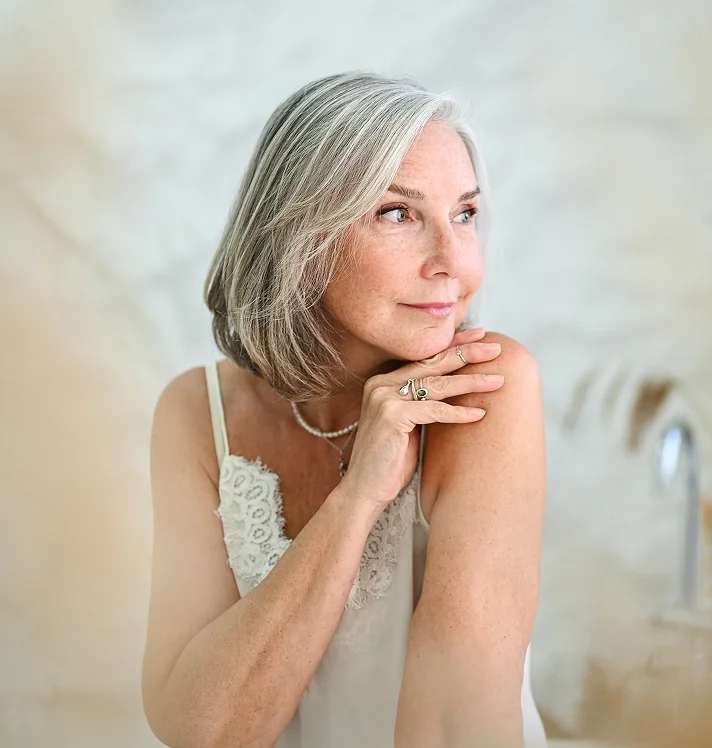 Mature woman with elegant gray hair and natural makeup in contemplative pose wearing white sleeveless top - reconstructive in San Diego, CA.