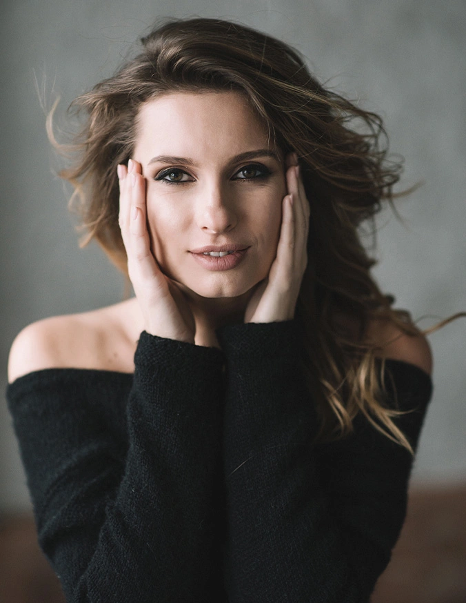 Woman in black off-shoulder top with flowing brown hair posing with hands near face against gray background - brow lift in San Diego, CA