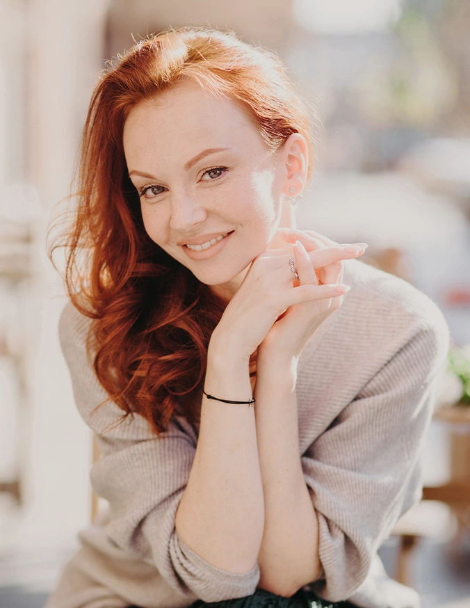 Smiling red-haired woman with hands clasped near face in soft natural lighting portrait - Chin augmentation in San Diego, CA
