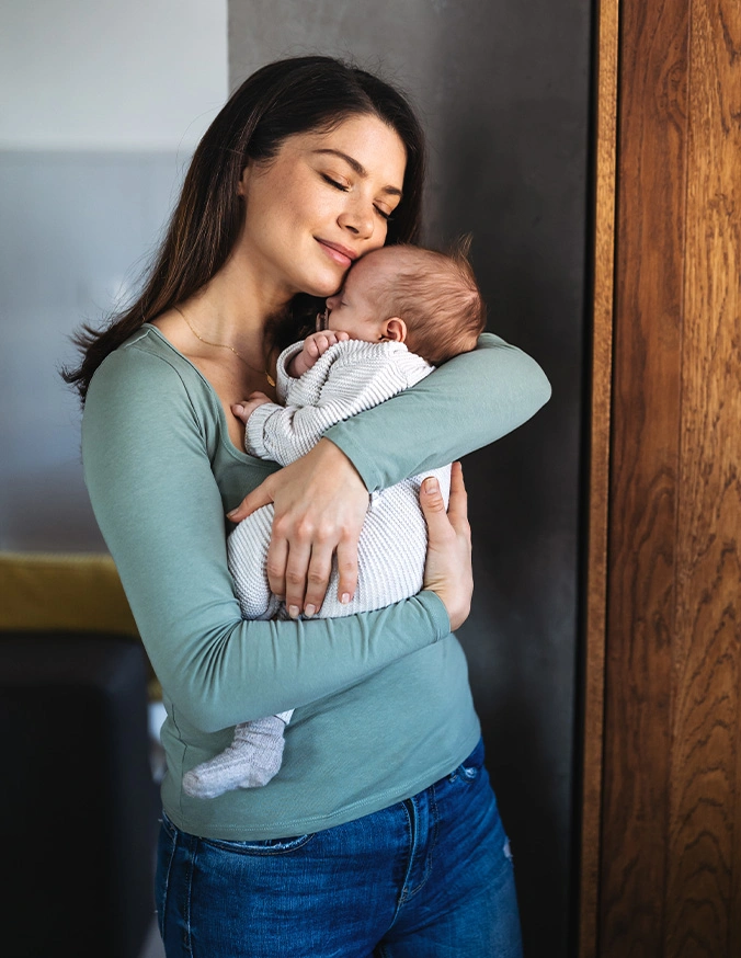 Mother lovingly holding and kissing newborn baby while standing indoors in casual clothing - ear reshaping in San Diego, CA