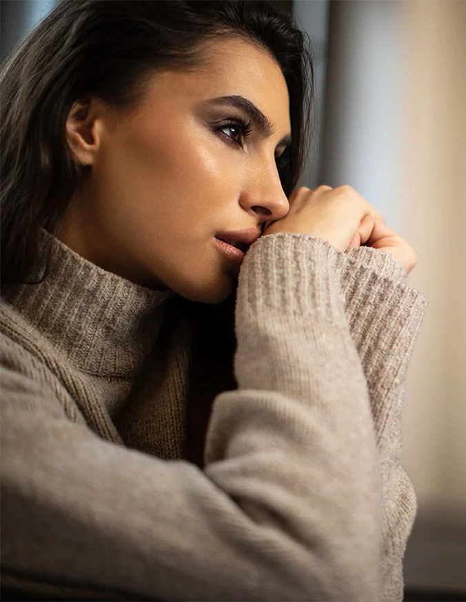 Woman in beige turtleneck sweater looking pensively out window in contemplative mood - ethnic rhinoplasty in San Diego, CA