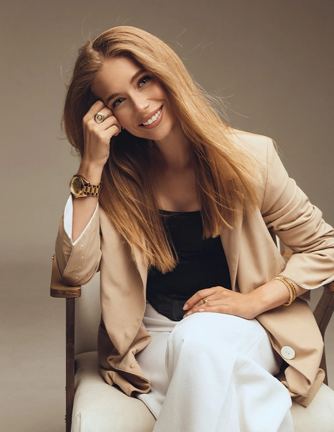 Professional woman in beige blazer and white pants smiling while sitting, business portrait on neutral background - facial fat grafting in San Diego, CA