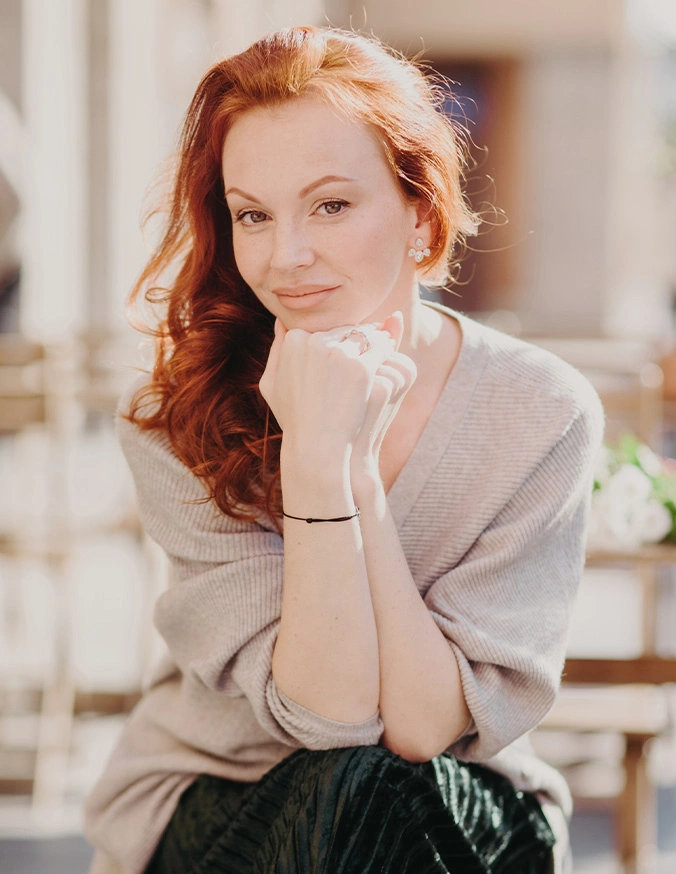 Red-haired woman in beige wrap and dark skirt posing with hands clasped near face in soft natural lighting - mole removal in San Diego, CA